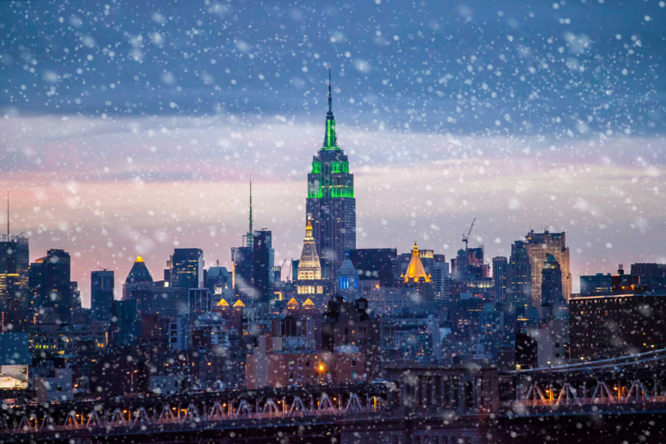 Snow falling over a city skyline at dusk, dominated by a tall building with green lights.