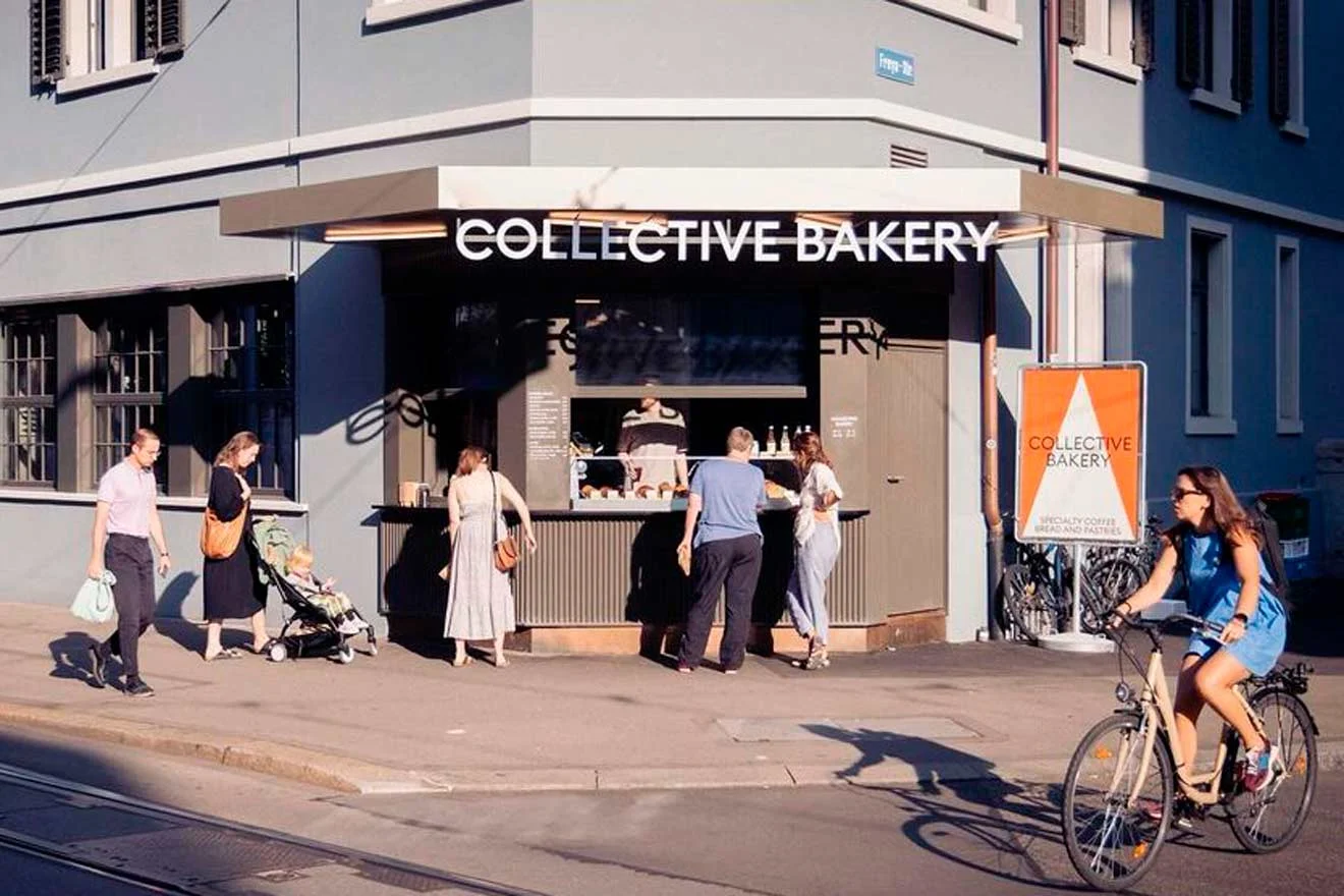 People stand outside Collective Bakery, with several interacting at the counter. A person rides a bike past. Buildings and a street sign are visible.