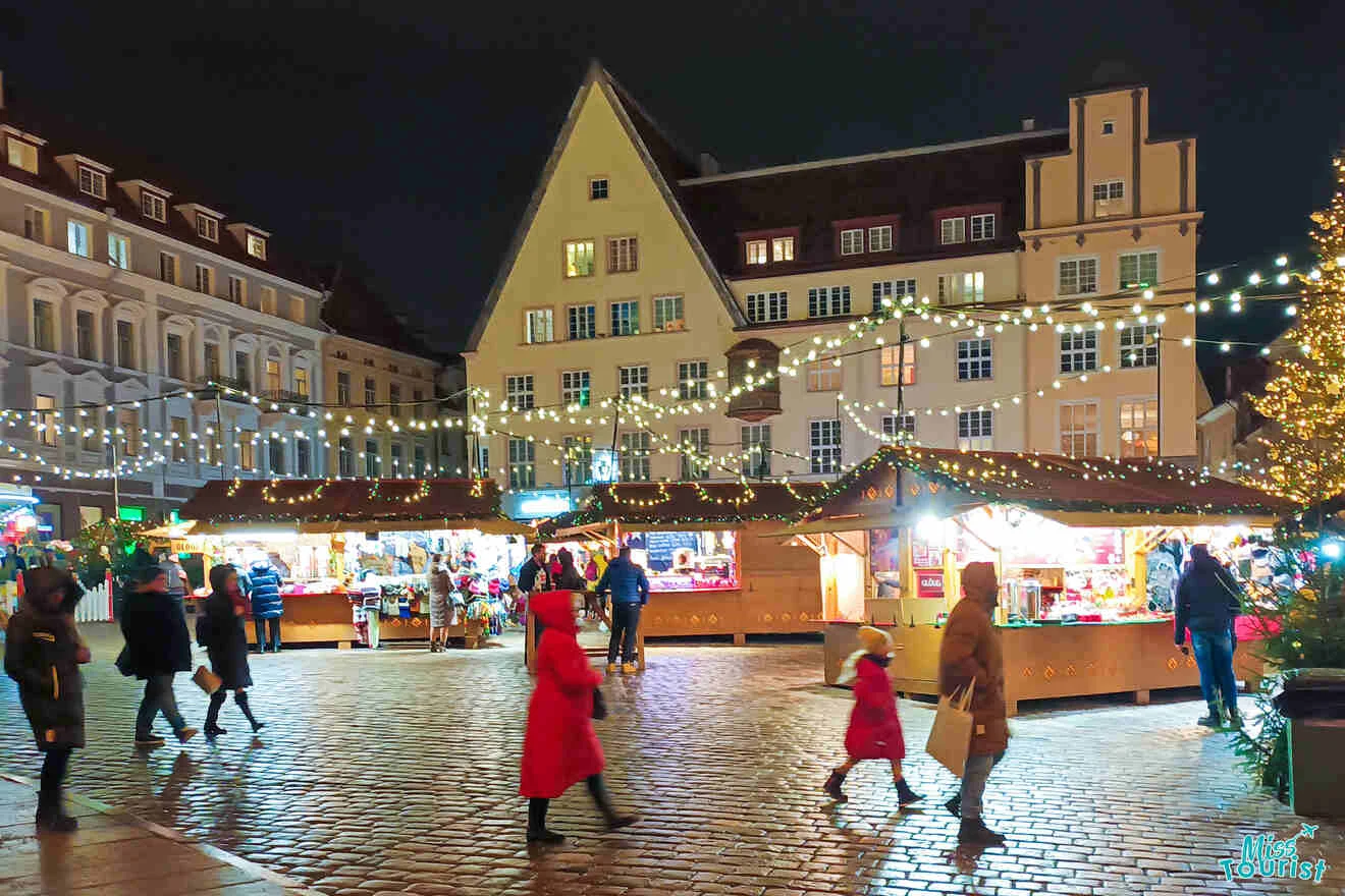 People walking in a festive market square at night, with wooden stalls, string lights, and illuminated buildings in the background.