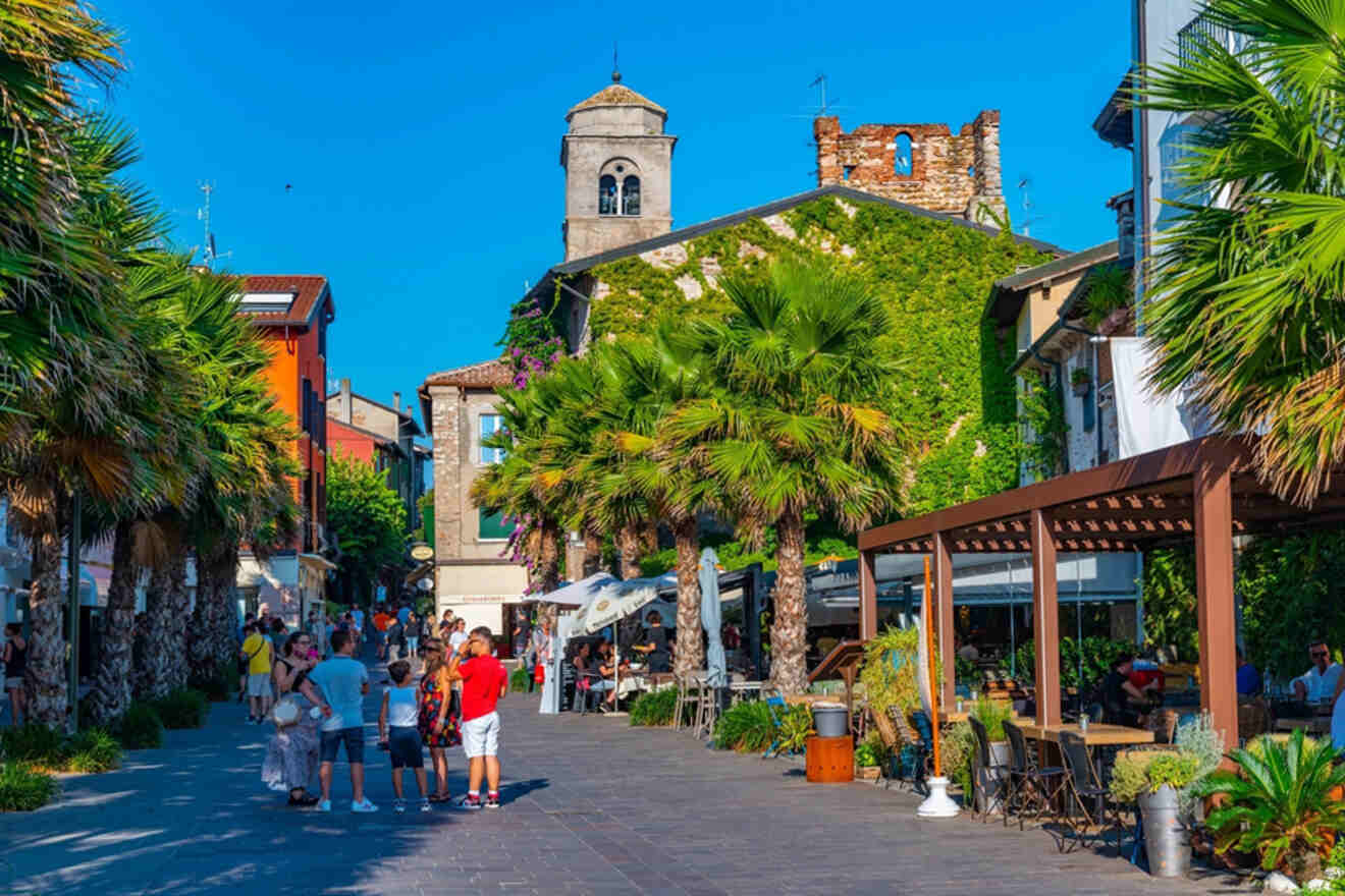 A vibrant street in a historic town features a group of people, palm trees, and a building with ivy-covered walls and a clock tower under a clear blue sky.