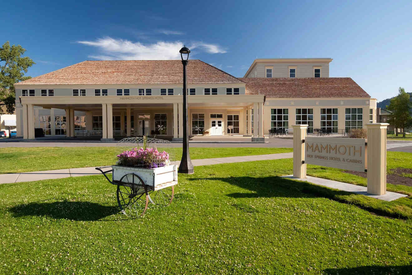 A building with a large entrance and a sign reading "MAMMOTH." A wooden cart with flowers is on the grass in front. Clear blue sky in the background.