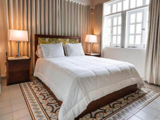 A cozy bedroom with a neatly made bed featuring white linens, flanked by two bedside tables with lamps, and a large window with curtains in the background.