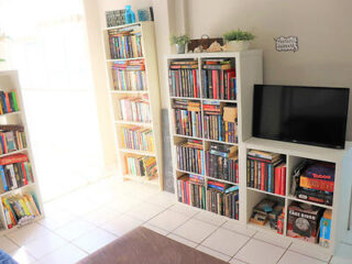 Room with bookshelves filled with books, a television, and board games on the lower shelf. The interior is brightly lit, with white walls and tiles.