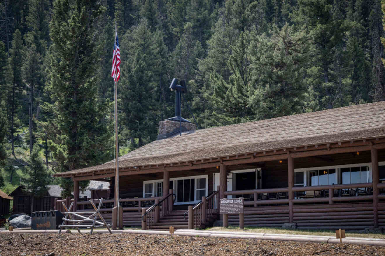 Wooden lodge with a stone chimney surrounded by tall trees, featuring an American flag on a pole and a wooden fence in the foreground.