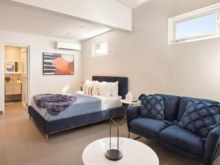 Minimalist bedroom with a navy bed, matching couch, decorative pillows, a round table, and a wall painting. An open door leads to a bathroom.