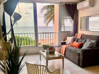 A cozy living room with a sofa, coffee table, and floor lamp, overlooking a balcony with a beach view.