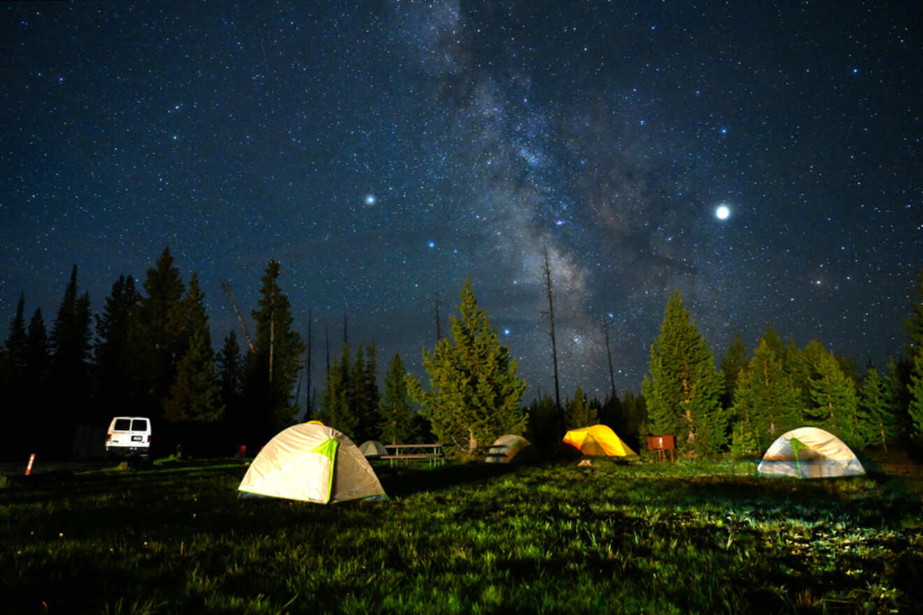 Several tents are set up in a grassy area under a starry night sky. The Milky Way is clearly visible above a pine forest, with a van parked nearby.