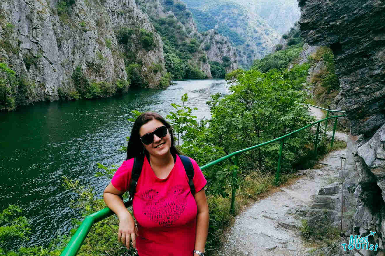 A person in a pink shirt and sunglasses stands on a riverside path surrounded by greenery and cliffs.