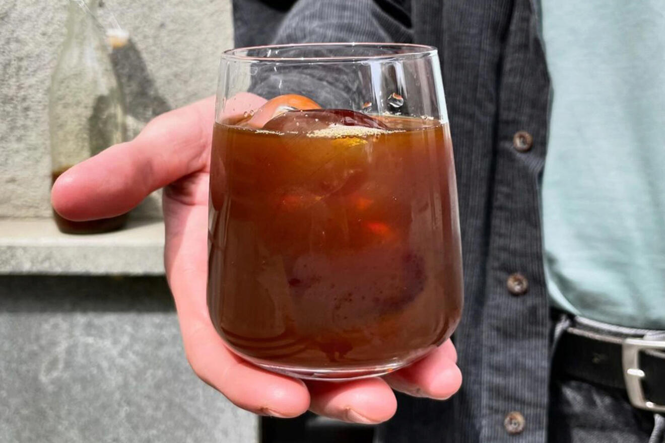 Person holding a glass of iced coffee close to the camera, with a wall and part of their shirt visible in the background.