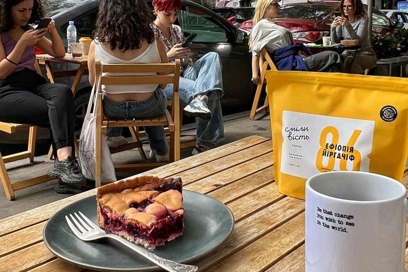 Outdoor cafe scene with several people seated. Focus on a slice of berry pie on a plate, a coffee cup, and a yellow bag of coffee on a wooden table.