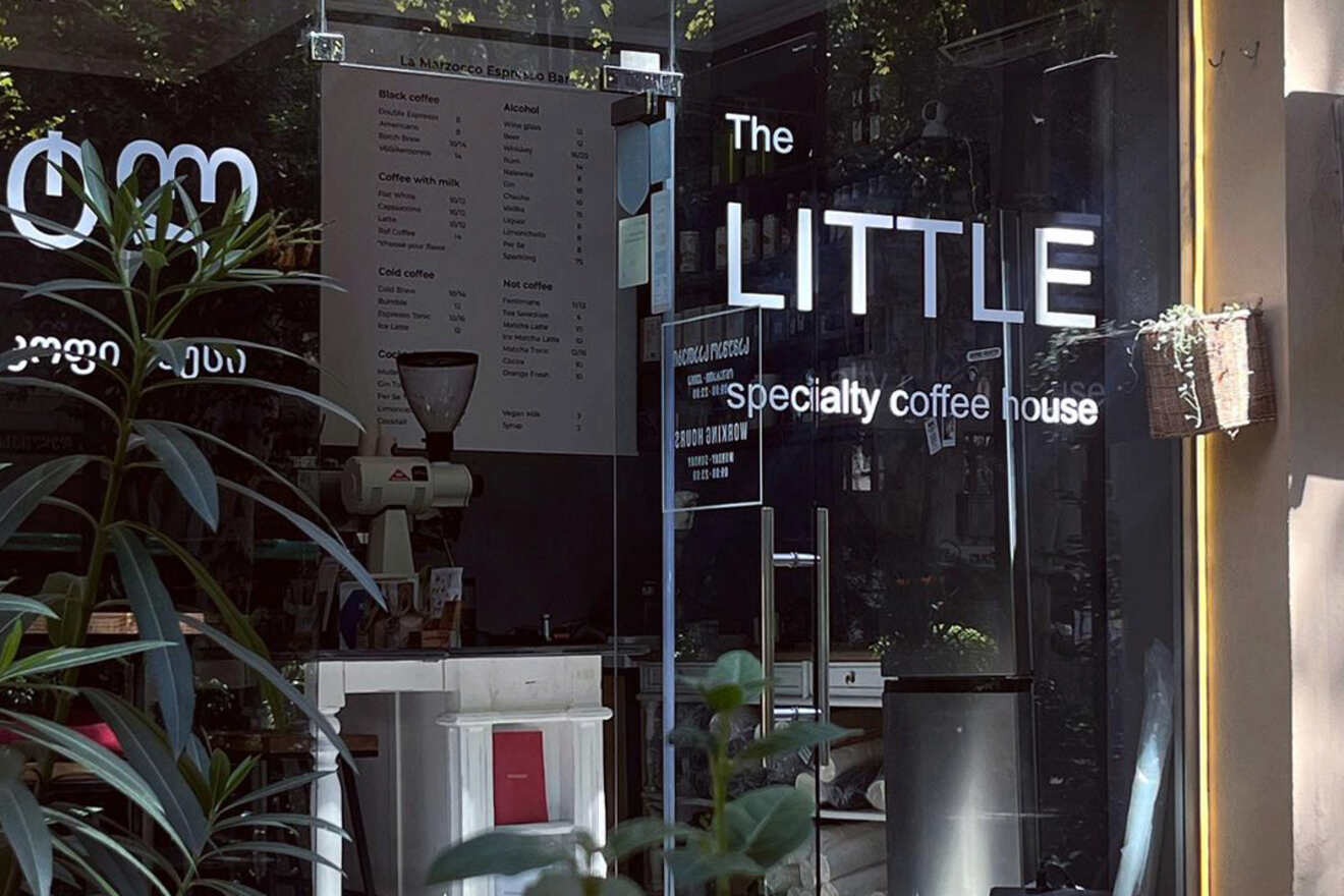 Front entrance of "The Little Specialty Coffee House" with menu board and a glimpse of the interior behind glass doors.