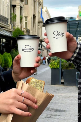 Two people holding coffee cups labeled "Stories" and "ზუნა & space." One holds a paper bag with a slice of bread or cake. They are outdoors on a cobblestone street with buildings in the background.