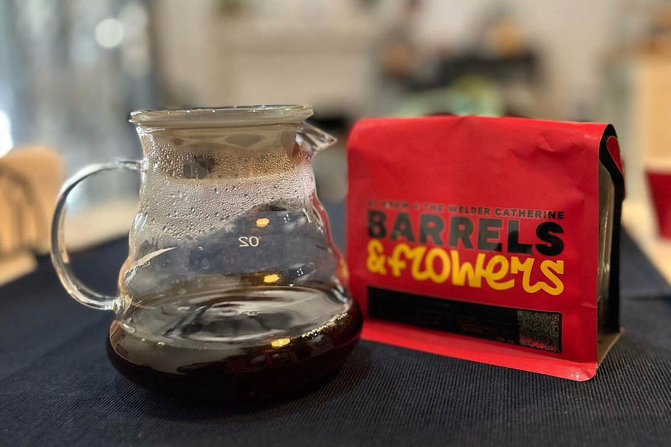 A glass coffee pot filled with dark liquid sits next to a red coffee bag labeled "Barrels & Flowers" on a dark tablecloth.