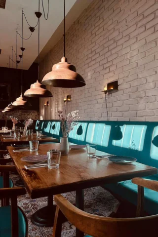 Restaurant interior with wooden tables, teal cushioned seating, and copper pendant lights. Tables set with plates and glasses, against a white brick wall.