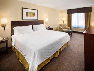 Hotel room with a king-sized bed, two nightstands, a lamp, a framed artwork above the bed, a chair with an ottoman, and a window with curtains.