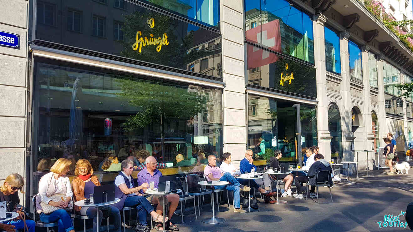 People sitting at outdoor tables of a café on a sunny day. Glass windows reflect city buildings, and a Swiss flag is visible.