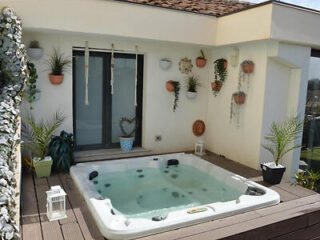 Outdoor jacuzzi on a wooden deck, surrounded by potted plants hanging on the wall. There's a sliding glass door and a lantern on the deck.