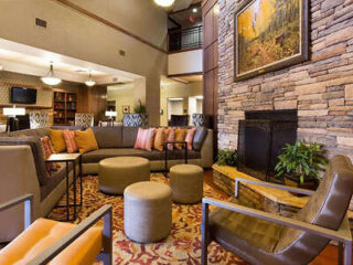 Cozy seating area with a large sectional sofa, armchairs, and round ottomans. A stone fireplace is on the right with a painting above it. The space has warm lighting and decorative plants.