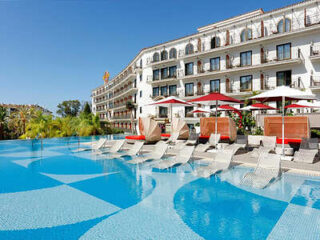 A luxurious hotel with a curved façade, featuring a large outdoor pool surrounded by lounge chairs and umbrellas, under a clear blue sky.