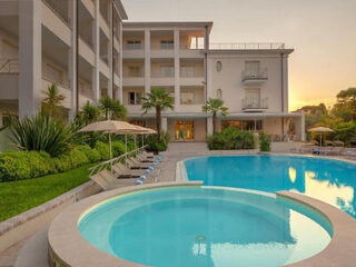 A modern hotel exterior with a round pool in the foreground and a larger pool surrounded by lounge chairs. The building is three stories high, with balconies and a sunset in the background.