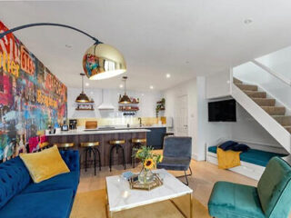 Modern living room with colorful graffiti wall, blue and yellow seating, a marble coffee table, and a nearby kitchen with bar stools. A staircase and mounted TV are in the background.