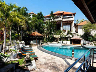 A resort with a pool surrounded by lounge chairs and tropical plants. The main building has multiple levels and red roofs, set against a clear blue sky.