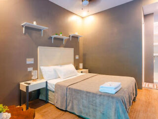 A neatly made bed with a gray cover in a modern bedroom featuring two bedside tables, a ceiling fan, and wall shelves with small decorative items.