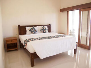 A simple bedroom with a wooden bed, white bedding, and patterned pillows. There is a bedside table and a sliding glass door leading outside.