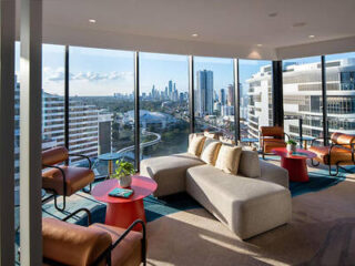 Modern lounge area with large windows offering a city skyline view. The room features a white sofa, red tables, and brown chairs.