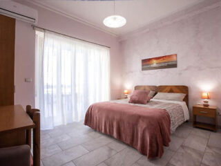 A bedroom with a double bed, pink bedspread, bedside tables with lamps, large window with sheer curtains, and a landscape painting on the wall.