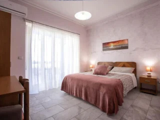 A bedroom with a double bed, pink bedspread, bedside tables with lamps, large window with sheer curtains, and a landscape painting on the wall.
