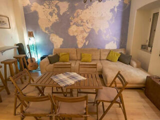 A cozy living room with a world map mural, beige sectional sofa, green and yellow pillows, wooden chairs, and a guitar in the corner.