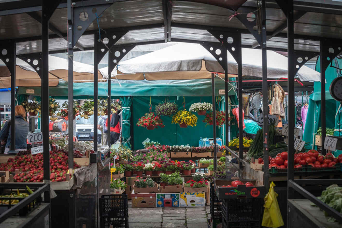 A variety of fruits, vegetables, and flowers are displayed at a covered outdoor market stall.