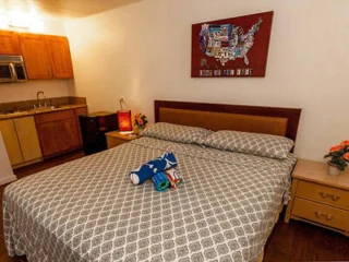 A small bedroom with a king-sized bed, a kitchenette in the corner, and a wall map decor. A bedside table holds a flower vase, and a towel and toiletries are placed on the bed.