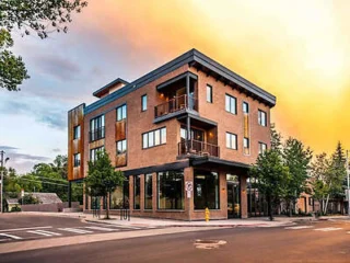 Three-story modern brick building with large windows on a street corner. Trees line the sidewalks, and the sky is a vivid orange.
