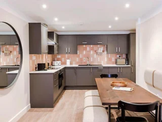 A modern kitchen with dark cabinets, pink-tiled backsplash, and wooden table with chairs. A large round mirror is on the wall, and ceiling lights are illuminated.