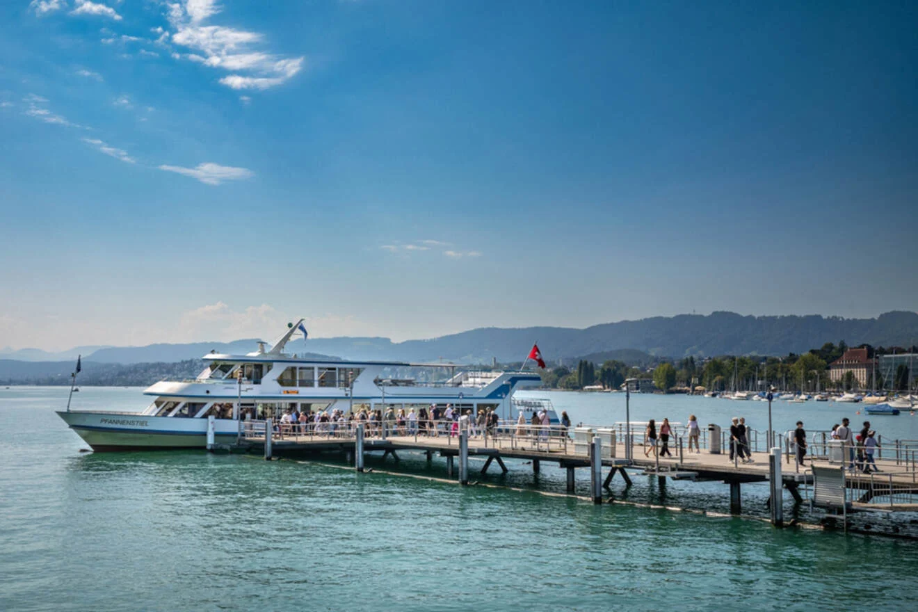 A passenger ferry is docked at a pier on a lake, with people boarding. The scene includes surrounding hills and a clear blue sky.