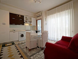 A modern, compact kitchen and dining area with white cabinets, a washing machine, glass table, red sofa, and large window with sheer curtains.