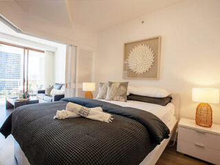 Contemporary bedroom with a neatly made bed, decorative pillows, two bedside lamps, and wall art. A view of an adjacent living area with large windows is visible.