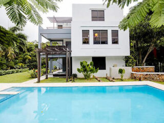 Modern white house with large windows, surrounded by greenery. It features a swimming pool in the foreground and a small patio area with a pergola.