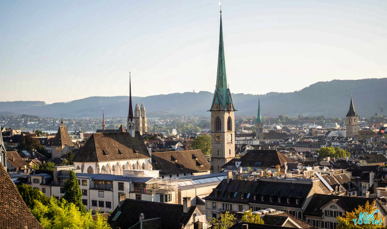 Skyline of a city featuring a tall church spire, surrounded by historic buildings with a hilly backdrop under a clear sky.