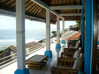 A shaded terrace with wooden furniture overlooks the ocean, with rooftops and sea visible in the background.