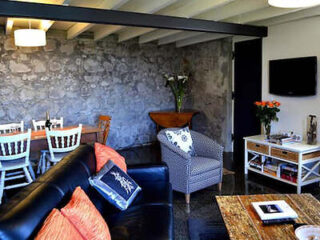 A cozy living room with a black sofa, decorative pillows, a patterned armchair, wall-mounted TV, wooden dining table with chairs, stone accent wall, and ceiling beams. Orange flowers on a side table.