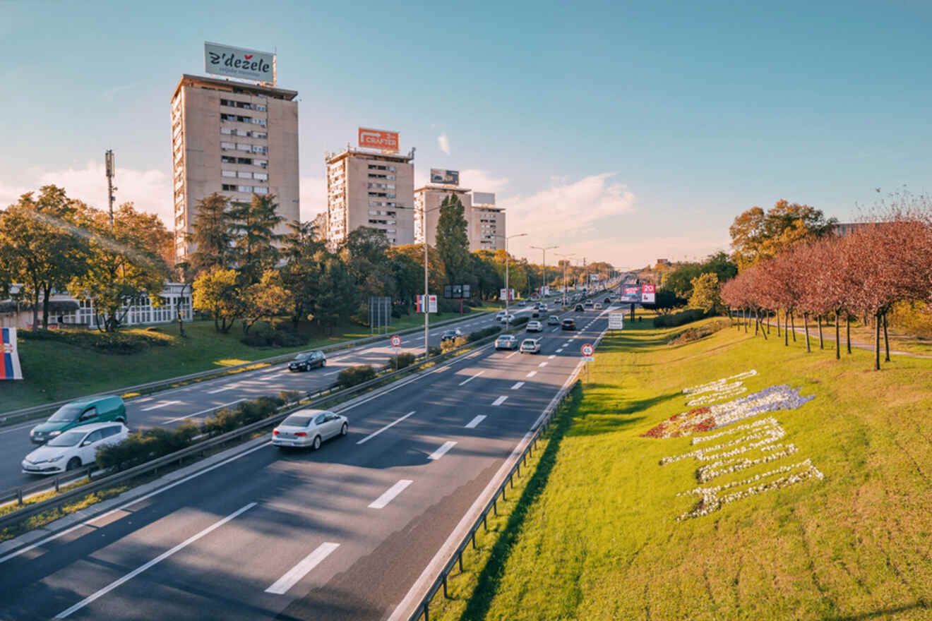 A busy urban road with multiple vehicles is flanked by grassy areas, trees, and tall buildings under a clear blue sky.