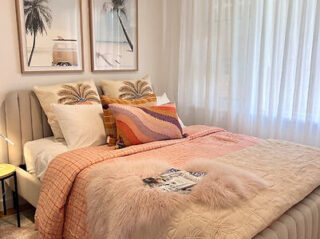 A cozy bedroom with a neatly made bed featuring pink and beige blankets, decorative pillows, and wall art of palm trees. Soft light filters through sheer curtains.