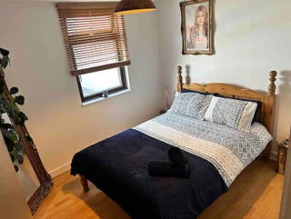 A small bedroom with a wooden bed, patterned bedding, a rolled black towel, a large mirror, a framed photo on the wall, wooden blinds, and a plant in the corner.