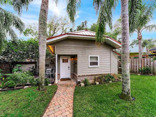 Small gray shed with a red roof, white door, and palm trees in front, set in a grassy yard with a brick pathway leading to the entrance.