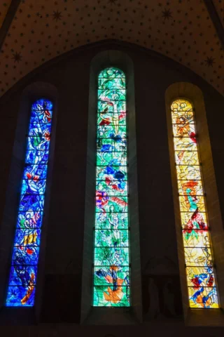 Three tall, narrow stained glass windows with abstract designs in blue, green, and yellow tones, set in a dimly lit arched space.