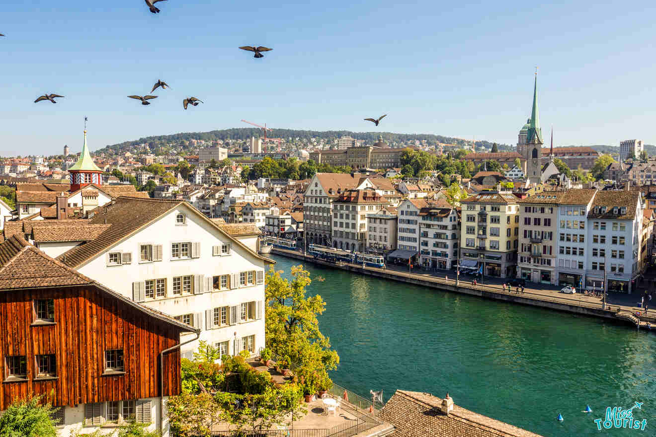 Aerial view of Zurich with old buildings, a river, and flying birds under a clear blue sky.