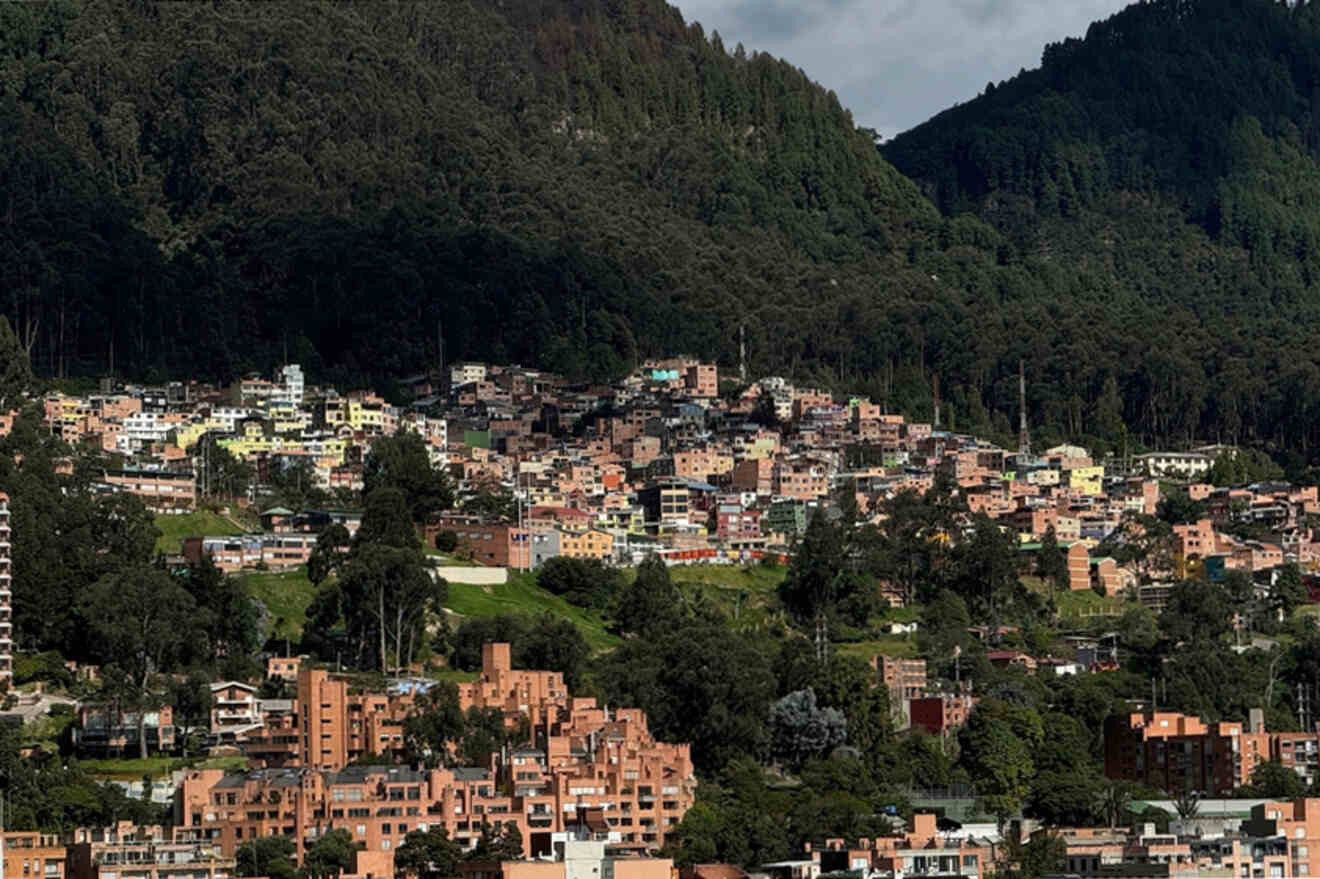 A hillside urban area with densely clustered buildings surrounded by green forests under a partly cloudy sky.